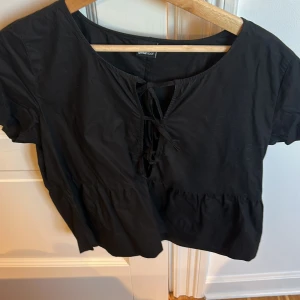 Svart kortärmad blus från Gina tricot - Snygg svart blus från Gina tricot med korta ärmar och knytning framtill. Perfekt för en stilren look. Blusen har en lös passform och är tillverkad i ett lätt material.