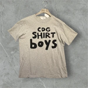 Grå t-shirt med tryck från CDG - Snygg grå t-shirt från CDG med svart texttryck 'CDG SHIRT BOYS' på framsidan.  Längd: 67 cm, bredd: 50 cm. Finns på folkungagatan 142!