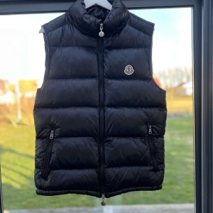 Svart dunväst från Moncler - Snygg svart dunväst från Moncler med dragkedja och hög krage. Västen har en quiltad design och praktiska fickor med dragkedja. Perfekt för kyligare dagar när du vill hålla stilen. Inga skador eller hål