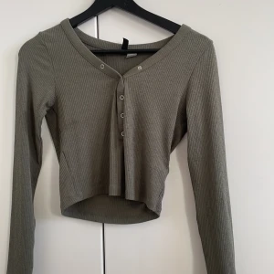 Olivgrön ribbad tröja - Säljer en olivgrön ribbad tröja med v-ringning och knappar framtill. Tröjan har långa ärmar och en croppad passform, perfekt för en avslappnad stil. Passar bra till både jeans och kjol.