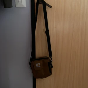 Brun axelväska från Carhartt - Säljer en snygg brun axelväska från Carhartt. Väskan har en justerbar axelrem och flera praktiska fack, inklusive ett med dragkedja på framsidan. Perfekt för att bära med sig det viktigaste på ett stilrent sätt.