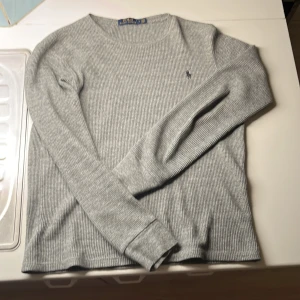 Grå ribbad tröja från Polo Ralph Lauren - Säljer en stilren grå ribbad tröja från Polo Ralph Lauren. Tröjan har långa ärmar och ett diskret broderat logotyp på bröstet. Perfekt för en avslappnad och snygg look.