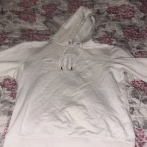 Vit hoodie från GANT - Säljer en stilren vit hoodie från GANT med broderad logga på framsidan. Hoodien har en justerbar dragsko i huvan och är perfekt för en avslappnad look. Passar bra till både jeans och joggers.