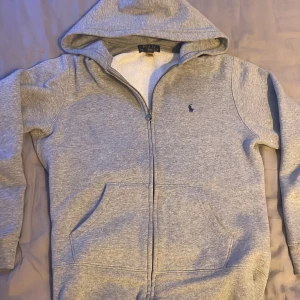 Grå hoodie från Ralph Lauren - Säljer en stilren grå hoodie från Ralph Lauren med dragkedja och huva. Fint begagnat skick. Storlek 14-16 / 160, men upplever storleken liten. Hör av dig vid intresse.
