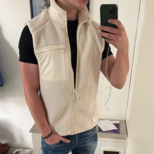 Beige fleeceväst från Only & Sons - Säljer en beige fleeceväst från Only & Sons. Västen har en dragkedja framtill och en praktisk bröstficka med dragkedja. Perfekt för lager-på-lager under kyligare dagar.