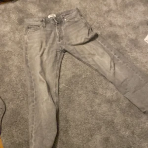 Grå jeans från Jack & Jones - Säljer ett par grå jeans från Jack & Jones med en relaxed passform. Modellen heter 'Relaxed/Chris' och har en klassisk design med fem fickor och knappgylf. Perfekta för en avslappnad stil.