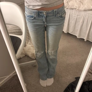 Ljusblå jeans med slitningar - Säljer ett par ljusblå jeans med slitningar på knäna. De har en klassisk femficksdesign och knappgylf. Perfekta för en avslappnad stil. Ett litet hål där vid bakfickan men inget som syns för mycket!