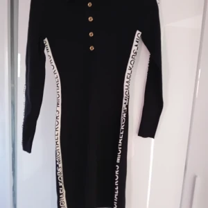Svart klänning från Michael Kors - Snygg svart klänning från Michael Kors med vita detaljer längs sidorna och knappar framtill. Klänningen har långa ärmar och en stilren design som passar perfekt för olika tillfällen.