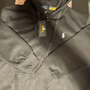 Polo Raulp lauren zip hoodie - Säljer en stilren svart hoodie från Polo Ralph Lauren med dragkedja och justerbar huva. Den har en broderad logga på bröstet och ribbade muddar vid ärmslut och nederkant. Perfekt för en avslappnad look.