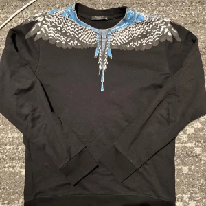 Marcelo Burlon - Säljer en svart tröja från Marcelo Burlon med ett coolt vingmönster i blått och grått runt halsen. Tröjan är långärmad och tillverkad i 100% bomull. Perfekt för dig som vill ha en unik stil! 🖤💙