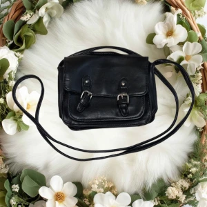 Svart axelväska - Snygg svart mindre axelväska i skinn med dubbla imitation spännen framtill. Väskan har en lång axelrem och en praktisk uppdelad ficka. Perfekt för att bära dina dagliga nödvändigheter med stil.