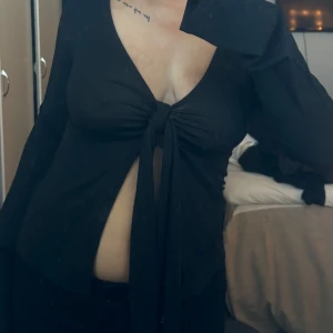 Svart långärmad knytblus - Säljer en elegant svart långärmad blus med knytning framtill. Perfekt för en stilren look. Blusen har en djup v-ringning och är tillverkad i ett mjukt material.