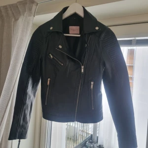 Svart skinnjacka från Livelo - Snygg svart skinnjacka från Livelo med dragkedjor och stilrena detaljer. Jackan har en klassisk bikerstil med långa ärmar och silverfärgade knappar.