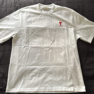 Vit t-shirt från Ami Paris - Säljer en stilren vit t-shirt från Ami med ett rött broderat hjärta och bokstaven 'A' på bröstet. Perfekt nu till sommaren när man vill ha en fin T-shirt. OBS ”bara testad aldrig använd”. Va inte rädd för att höra av dig vid frågor!