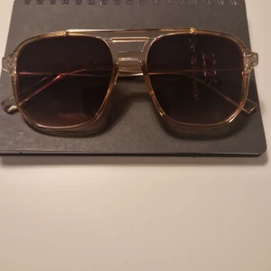 Transparenta solglasögon med brun toning - Snygga solglasögon med transparent båge och bruna tonade glas. De har en modern design med en dubbel brygga och breda skalmar. Perfekta för en stilren look.