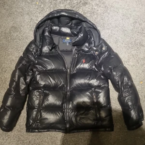 Svart pufferjacka från Polo Ralph Lauren - Säljer en snygg svart pufferjacka från Polo Ralph Lauren. Jackan har en quiltad design med en dragkedja framtill och en huva för extra värme. Den ikoniska röda loggan är broderad på bröstet. Perfekt för kyliga dagar!