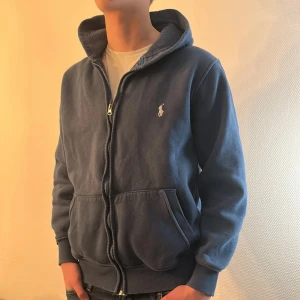 Blå hoodie från Ralph Lauren - Säljer en snygg blå hoodie från Ralph Lauren med dragkedja och broderad logga på bröstet. Perfekt för en avslappnad stil. Hoodien har långa ärmar och en bekväm passform.