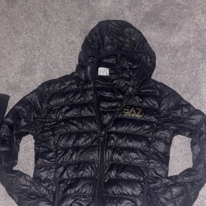 Svart dunjacka från Emporio Armani - Snygg svart dunjacka från Emporio Armani med EA7-logga i guld på bröstet. Jackan har jag använda jag i cirka en månad efter jag köpte den men sedan tröttna jag på den eftersom den ej är min stil.