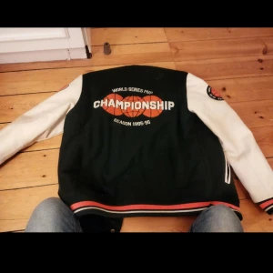 Svart och vit bomberjacka med broderier - Säljer en grön och vit bomberjacka med coola broderier och applikationer. Jackan har ett stort 'P' på framsidan och texten 'CHAMPIONSHIP' på baksidan. Ärmarna är vita med broderade märken som 'MVP' och 'World Series'. Perfekt för en sportig look!