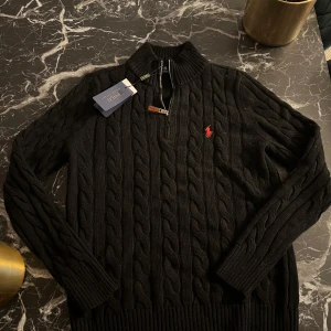 Svart kabelstickad tröja från Polo Ralph Lauren - Säljer en stilren svart kabelstickad tröja från Polo Ralph Lauren med en broderad röd logotyp på bröstet. Tröjan har en dragkedja vid halsen och långa ärmar. Perfekt för en klassisk look.