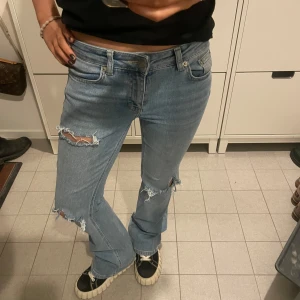 Blå jeans med slitningar - Säljer ett par blå jeans med coola slitningar på knäna. De har en klassisk femficksdesign och en bekväm passform. Perfekta för en avslappnad stil.