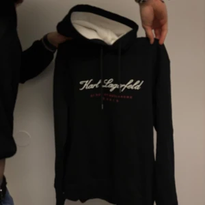 Svart hoodie från Karl Lagerfeld - Snygg svart hoodie från Karl Lagerfeld med vit text på bröstet. Den har en justerbar huva med vita snören och en mjuk insida. Perfekt för en stilren och bekväm look.