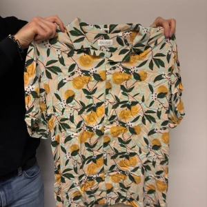 Blommig kortärmad skjorta från Urban Outfitters - Säljer en färgglad kortärmad skjorta från These Glory Days med ett blommigt mönster i gult och grönt. Perfekt för sommarens alla tillfällen. Skjortan har knappar framtill och en klassisk krage.