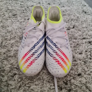 Adidas Predator fotbollsskor - Snygga Adidas Predator fotbollsskor i vitt med färgglada detaljer i blått, rött och gult. Skorna har snörning och en modern design som sticker ut på planen. Perfekta för den som vill ha stil och funktion i ett.