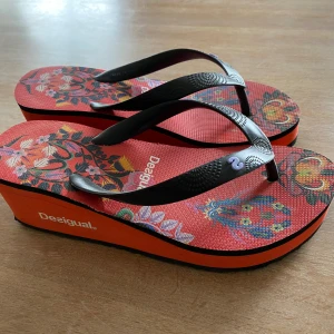 Skor från Desigual - Snygga flip-flops från Desigual med en färgglad blommig design på en röd bas. De har en kilklack och svarta remmar.
