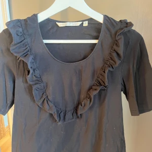 Svart topp med volang från House of Lola - Säljer en svart topp från House of Lola med korta ärmar och en fin volangdetalj runt halsringningen. Perfekt för en stilren look.