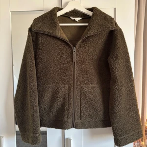 Teddyjacka - Mysig olivgrön teddyjacka med dragkedja framtill. Jackan har en avslappnad passform och långa ärmar, perfekt för kyliga dagar. Den har en enkel design med två fickor framtill och en hög krage för extra värme. Perfekt för höst och vinter!