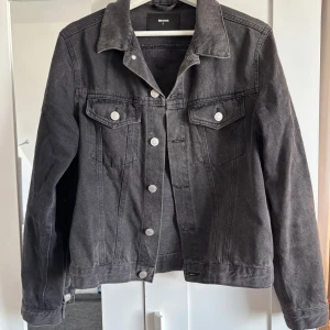 Jeansjacka  - Snygg svart jeansjacka från Bik Bok med klassisk design och silverfärgade knappar. Jackan har två bröstfickor med lock och knappar samt långa ärmar med knappslut. Perfekt för en casual look!