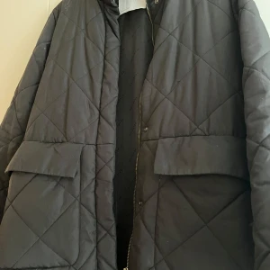 Svart quiltad jacka - Snygg svart quiltad jacka med dragkedja och knappar. Jackan har långa ärmar och stora fickor framtill. Perfekt för kyligare dagar.