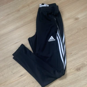 Svarta träningsbyxor från Adidas - Snygga svarta träningsbyxor från Adidas med de klassiska tre vita ränderna längs sidorna. Byxorna har en elastisk midja och är perfekta för sport och träning. Adidas-loggan är tryckt på ena benet.