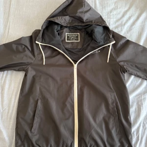 McKenzie vindjacka - Säljer en stilren svart vindjacka från McKenzie. Jackan har en praktisk huva och dragkedja framtill. Perfekt för blåsiga dagar och har en sportig look. Den är långärmad och har en enkel design utan mönster.