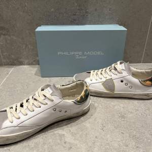 Snygga vita sneakers från Philippe Model med grå mockadetaljer och kamouflagemönster på hälen. Skorna har snörning och en stilren design med en touch av färg. Perfekta för en avslappnad stil. Skorna köpta förra veckan. Använda 4-5 gånger. Phillipe Model Junior så sitter normal i storlek.  Allt OG ingår. Nypris typ 3400kr. Tveka inte på att fråga! /Vikram