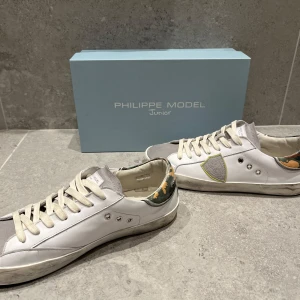 Phillipe Model skor 38 - Snygga vita sneakers från Philippe Model med grå mockadetaljer och kamouflagemönster på hälen. Skorna har snörning och en stilren design med en touch av färg. Perfekta för en avslappnad stil. Skorna köpta förra veckan. Använda 4-5 gånger. Phillipe Model Junior så sitter normal i storlek.  Allt OG ingår. Nypris typ 3400kr. Tveka inte på att fråga! /Vikram