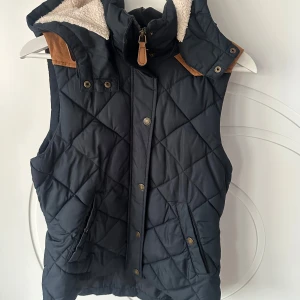 Quiltad väst från H&M - Snygg quiltad väst från H&M i mörkblått med bruna detaljer. Västen har en avtagbar huva och stängs med både dragkedja och knappar. Insidan är fodrad med ett rutigt mönster för extra stilpoäng. Perfekt för kyliga dagar!