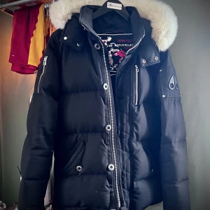 Svart dunjacka från Moose Knuckles - Snygg svart dunjacka från Moose Knuckles med vit päls och svart klövar på armemblemet. Bytes eventuellt mot Parajumper eller annan jacka, helst inte lika varm som Moose. Nypris- 13950:-