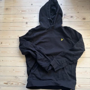 Svart hoodie från Lyle & Scott - Säljer en stilren svart hoodie från Lyle & Scott med deras ikoniska logga på bröstet. Tröjan har en justerbar huva och ribbade muddar vid ärmslut och nederkant. Perfekt för en avslappnad look.