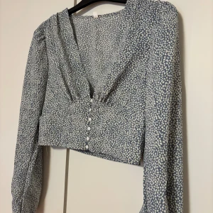 Blåmönstrad blus med knappar - Säljer en söt blåmönstrad blus med vita prickar och knappar framtill. Blusen har en v-ringad design och långa ärmar. 🩵