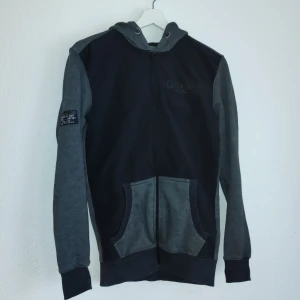 Grå och svart hoodie från F&F - Säljer en snygg grå och svart hoodie från F&F. Den har en dragkedja framtill och en praktisk ficka. Perfekt för en avslappnad stil. Huvan ger extra komfort och värme.