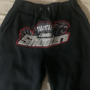 Svarta mjukisbyxor från Trapstar - Svarta mjukisbyxor från Trapstar med ett stort tryck på framsidan som säger 'Shooters'. Byxorna har en elastisk midja med dragsko och är perfekta för en avslappnad stil.