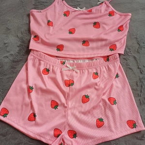 Rosa ribbad pyjamas med jordgubbsmönster - Söt rosa ribbad pyjamas med ett lekfullt jordgubbsmönster. Toppen har smala axelband och en liten rosett framtill som ger en extra charmig touch. Perfekt för en avslappnad och somrig look.