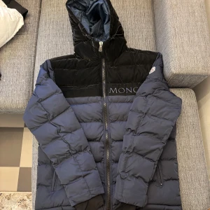 Svart och blå pufferjacka från Moncler - Snygg svart och blå pufferjacka från Moncler med huva och dragkedja. Jackan har långa ärmar och är perfekt för kyligare väder. Den har en quiltad design och praktiska fickor med dragkedja.