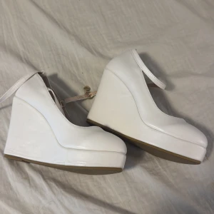 Vita kilklackar med rem - Snygga vita kilklackar med justerbar rem runt ankeln. Perfekta för att ge lite extra höjd och stil. De har en rund tå och en stabil kilklack som gör dem bekväma att bära.