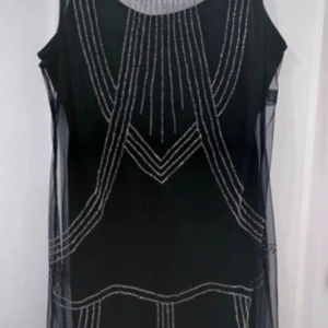 Svart ärmlös klänning från Bodyflirt - Snygg svart ärmlös klänning från Bodyflirt med silverfärgade detaljer i ett geometriskt mönster. Perfekt för en elegant kvällslook. Klänningen har en transparent överdel som ger en stilren touch.