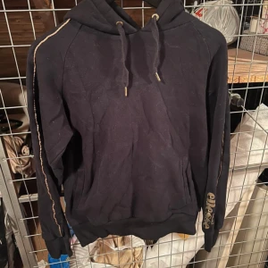 Svart hoodie från Ellesse - Säljer en svart hoodie från Ellesse med gulddetaljer och logga på armen. 
