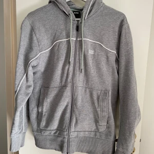 Hugo Boss grå tracksuit - Säljer en stilren grå tracksuit från Hugo Boss med dragkedja och justerbar huva. Tröjan har ribbade muddar och fickor framtill. Perfekt för en avslappnad look. Säljs då den inte används längre  och är dessutom i nyskick.