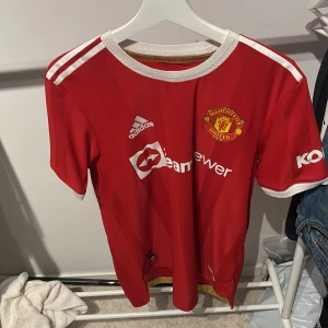 Manchester United Elanga 36 matchtröja - Röd Manchester United fotbollströja från Adidas med Elanga och nummer 36 på ryggen. Tröjan har vita detaljer på axlar och ärmslut samt klubbmärke på bröstet. Perfekt för fans som vill visa sitt stöd på läktaren eller hemma.